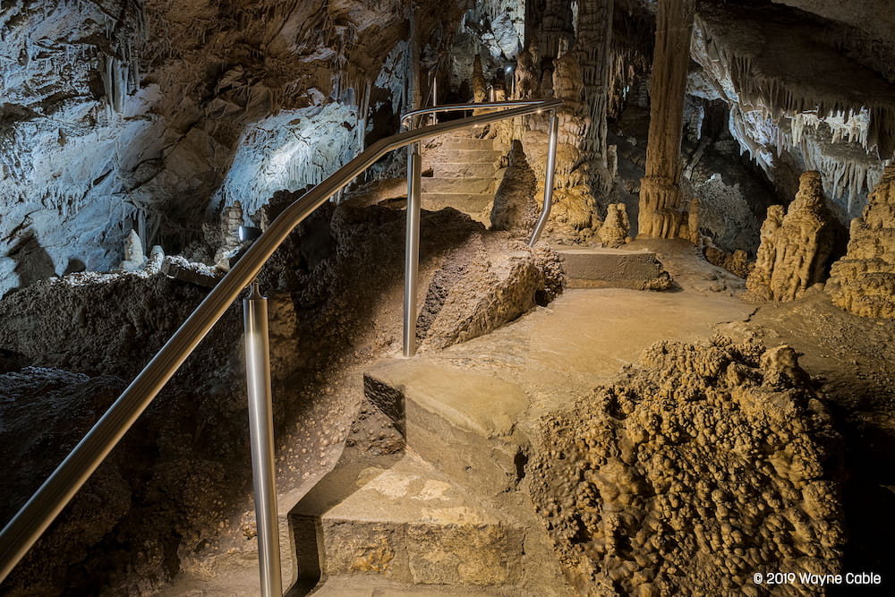 A dimly lit cave pathway with metal railings, surrounded by jagged stalactites and stalagmites. The atmosphere is mysterious and serene.