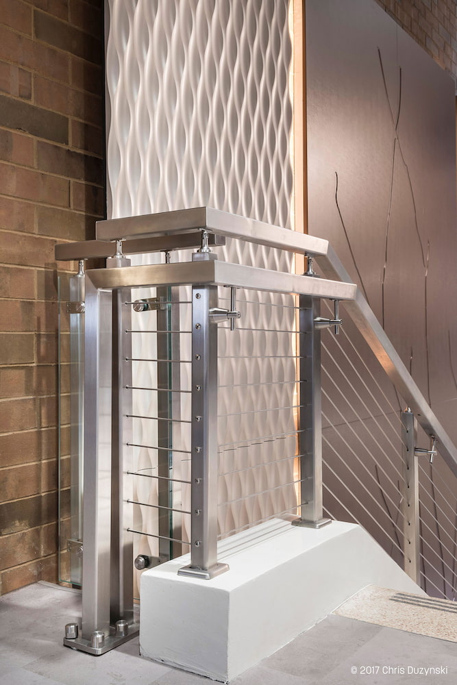 Modern staircase with metal railings, textured white wall, and a brown backdrop with linear patterns. The scene has a sleek, contemporary feel.