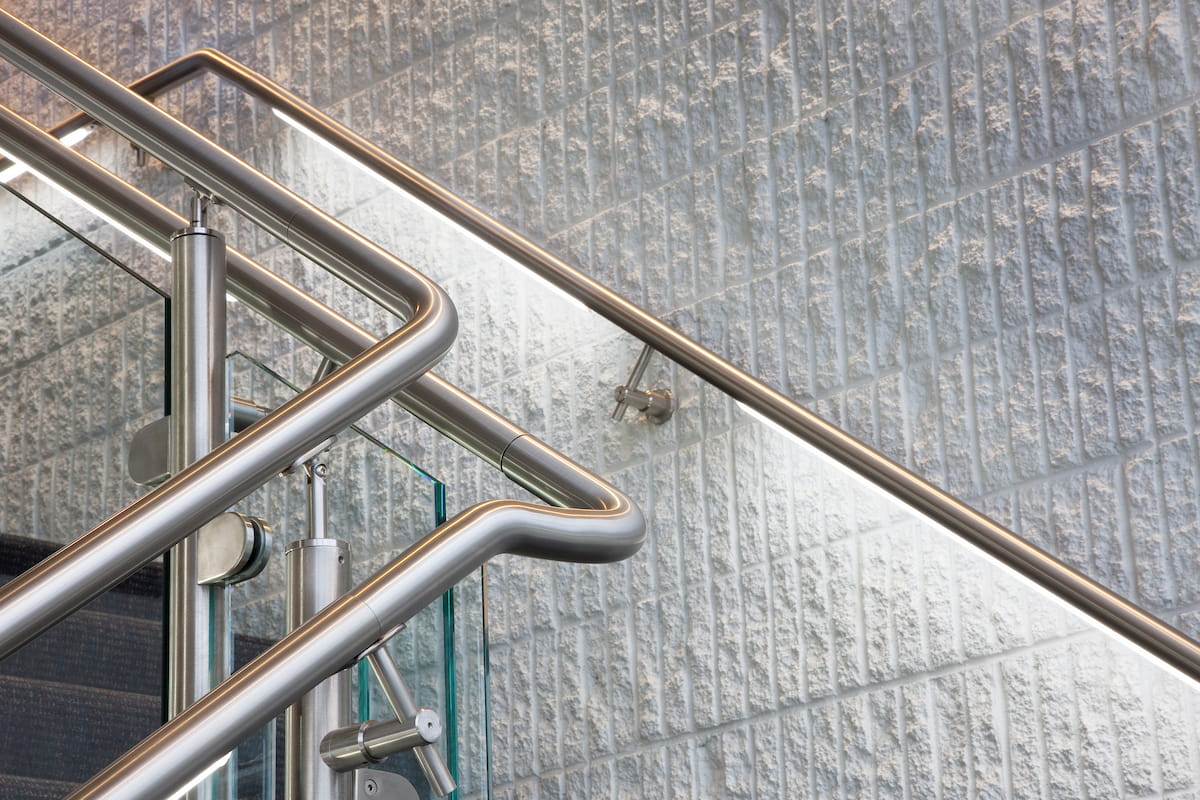 Modern stainless-steel stair railings curve against a textured glass-block wall, creating geometric lines and a sleek, industrial atmosphere.
