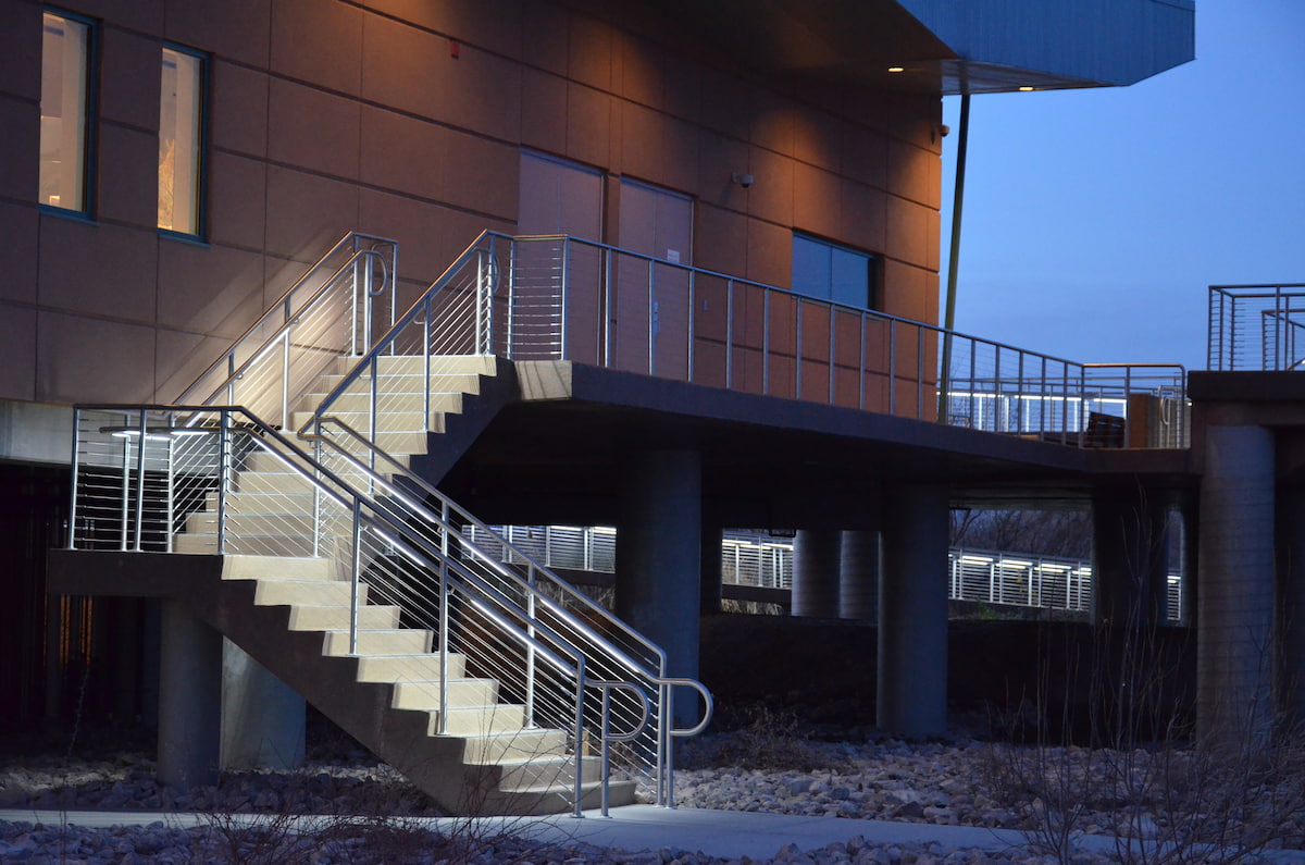 Modern building exterior with illuminated metal staircases, railings, and an elevated walkway at dusk; wintry ground creates a quiet mood.