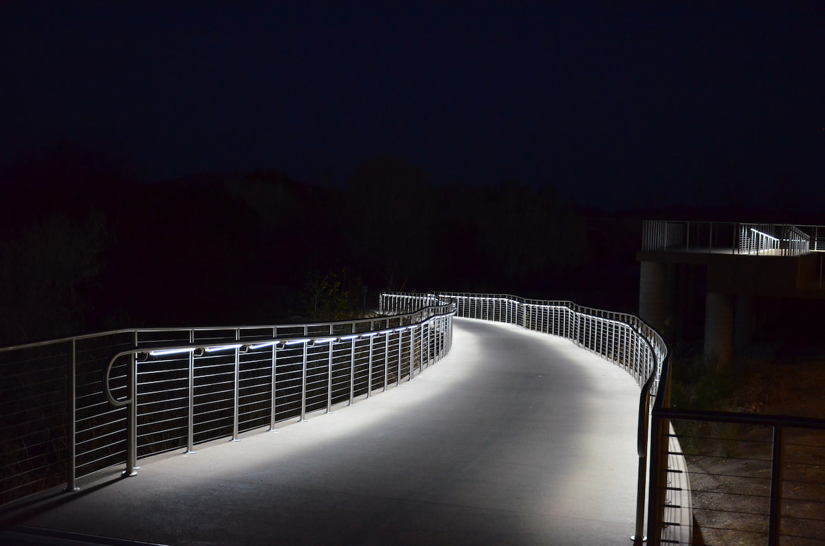Illuminated pedestrian bridge curves at night, with sleek metal railings and a viewing platform creating a serene mood.