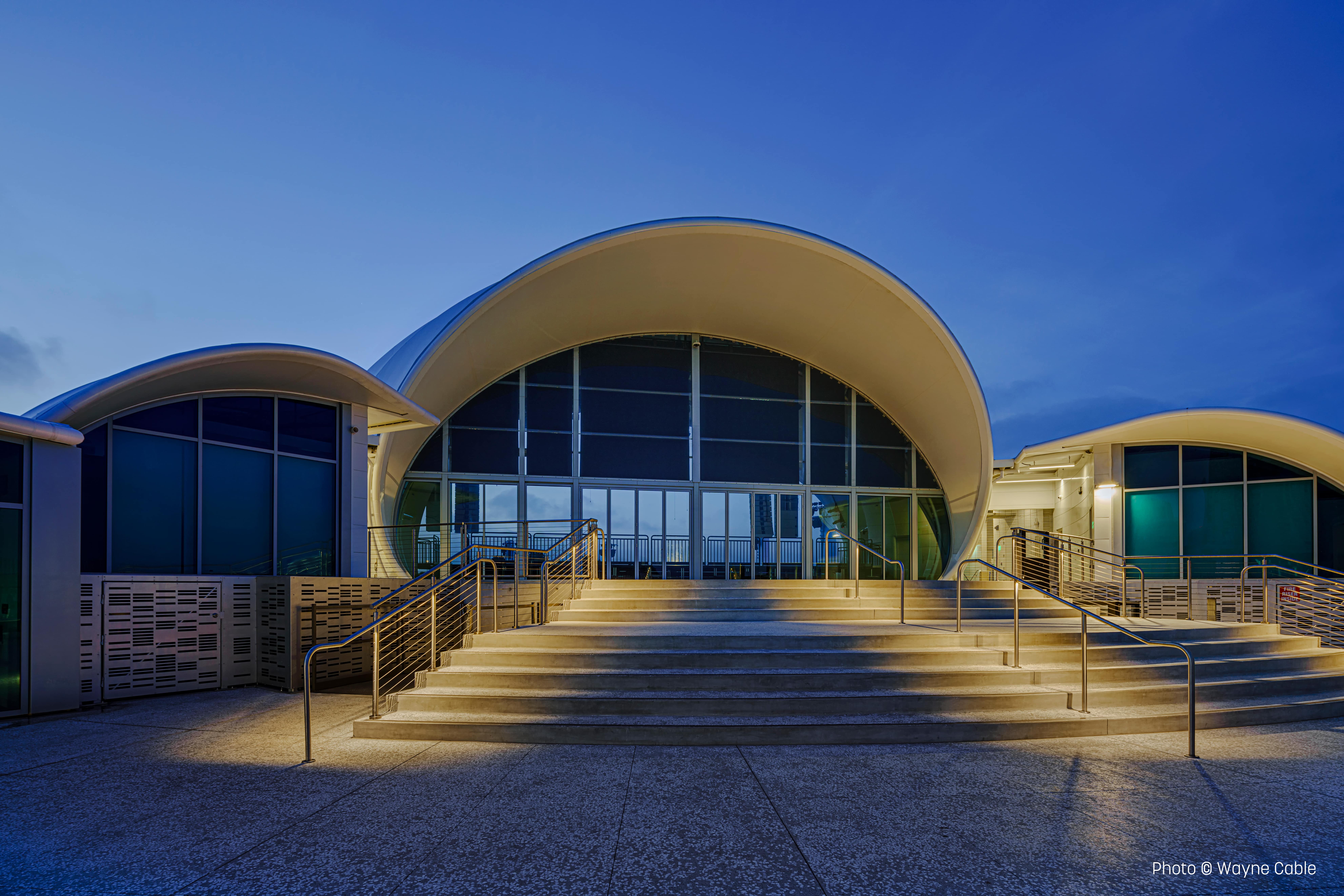 Twilight view of The Rady Shell at Jacobs Park, San Diego, with arched roof and illuminated steps leading to the glass entrance.