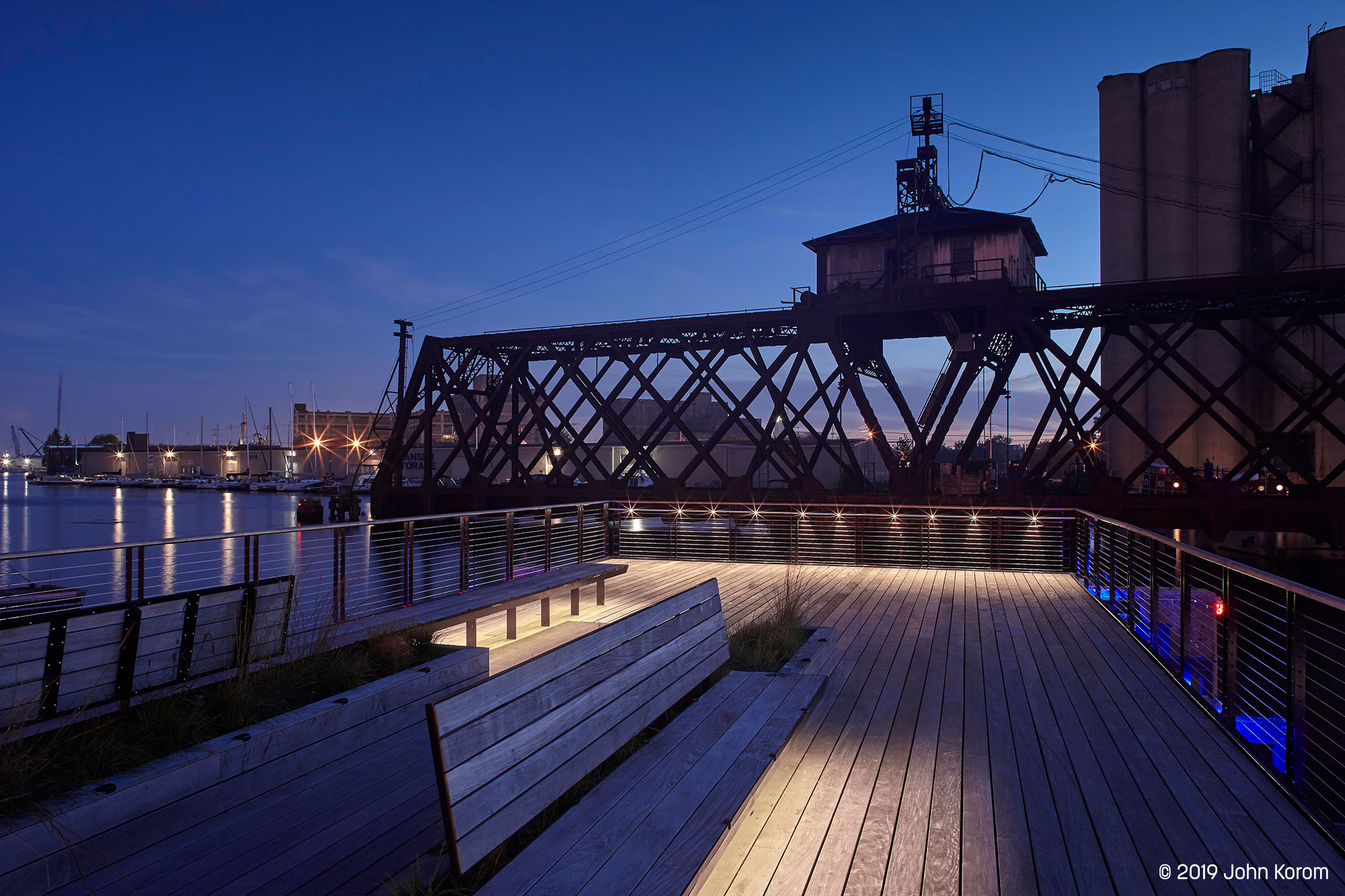 Trestle Park on Milwaukee Riverwalk at dusk, featuring Lumenpod 28 lighting, benches, and a historic train trestle bridge.