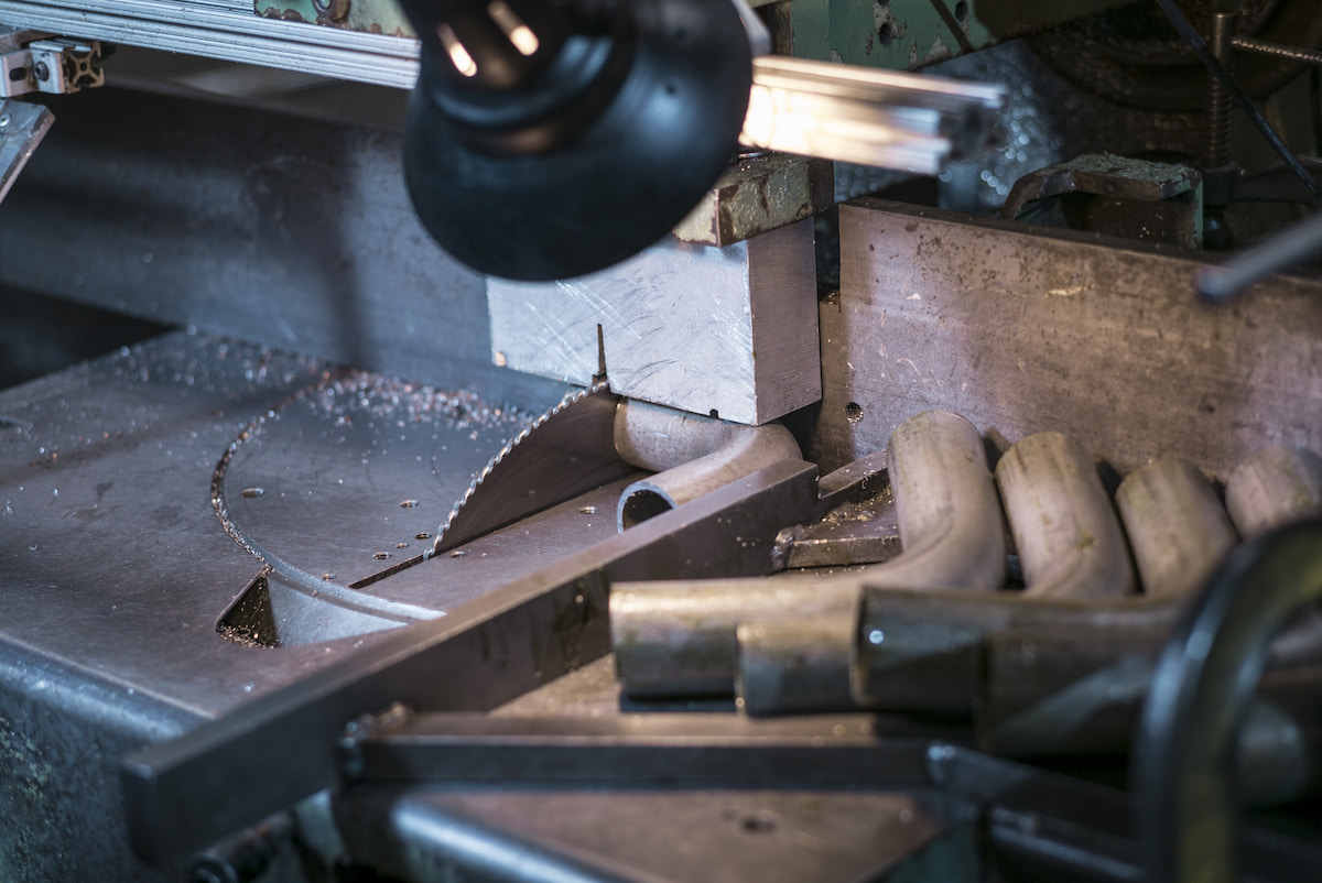 Close-up of an industrial metal-cutting machine with a circular saw blade, metal bars, and scattered shavings; gritty workshop atmosphere.