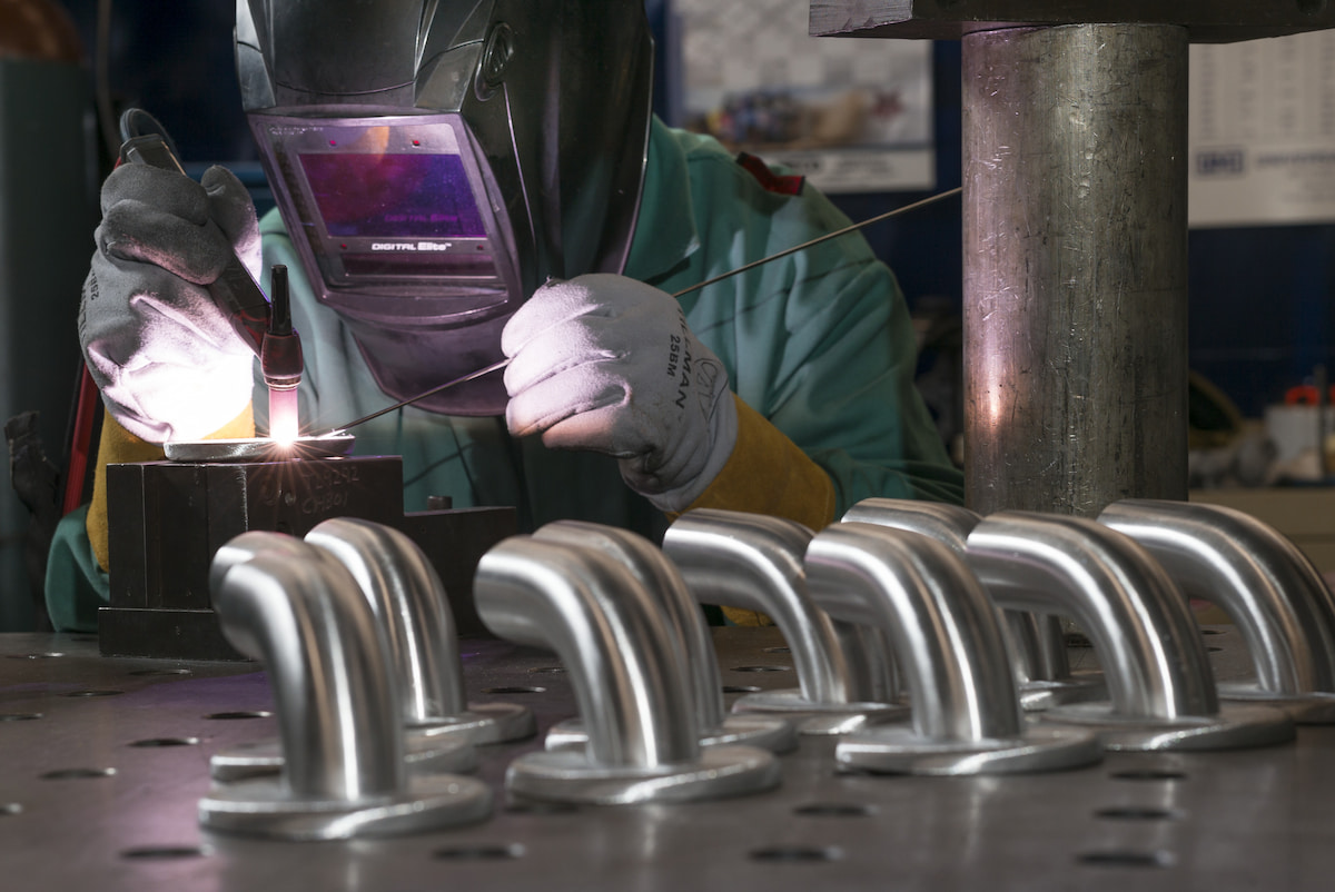 Welder in a protective helmet and gloves joins metal pipes, bright sparks glowing on a workshop table; rows of polished bends fill the foreground.