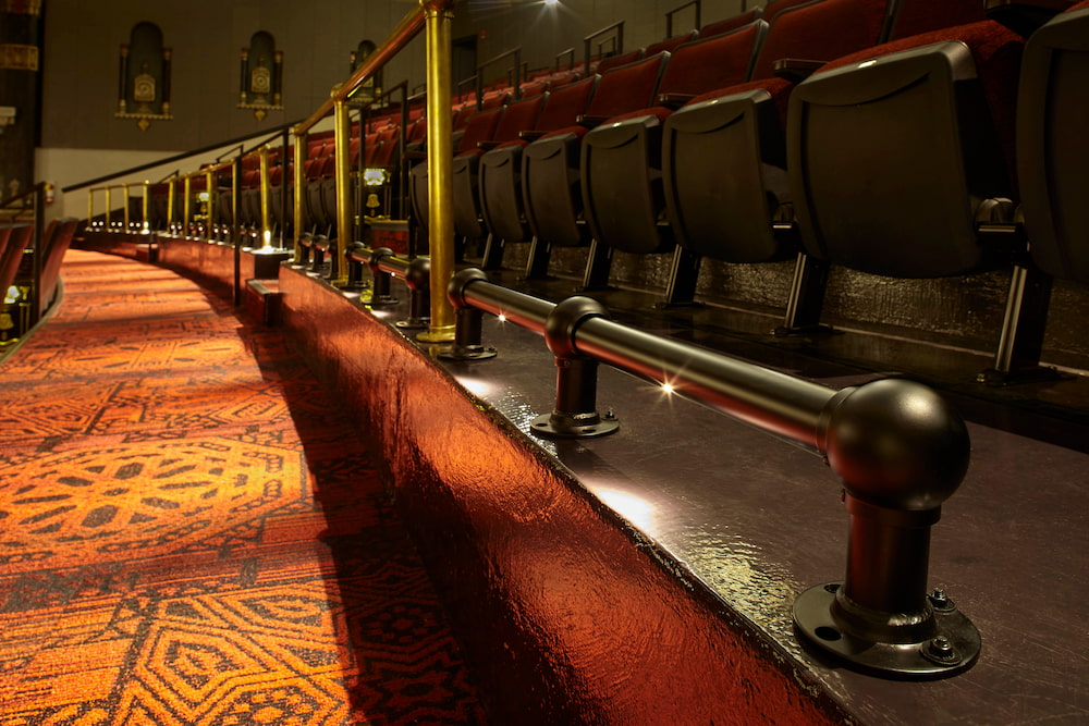 Close-up of theater seating with ornate red carpet and illuminated black foot railings in front.